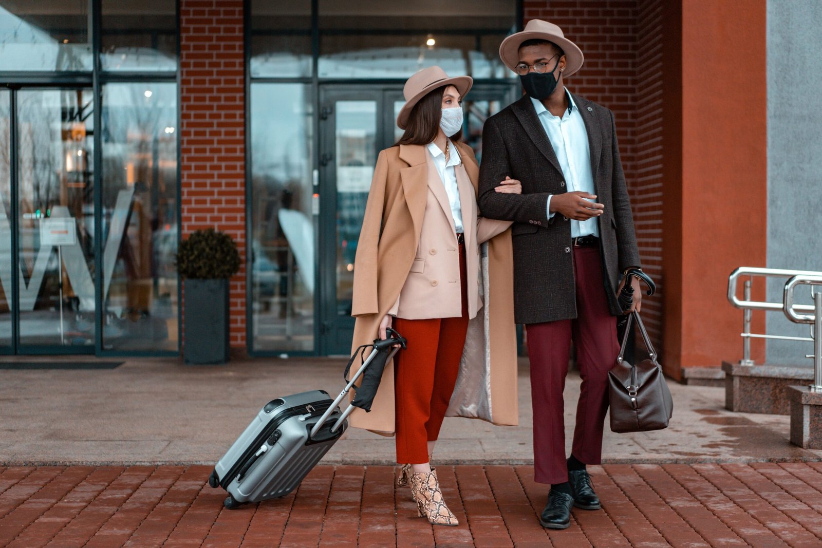 Home Fashionable couple with face masks at hotel entrance, showcasing travel during pandemic.